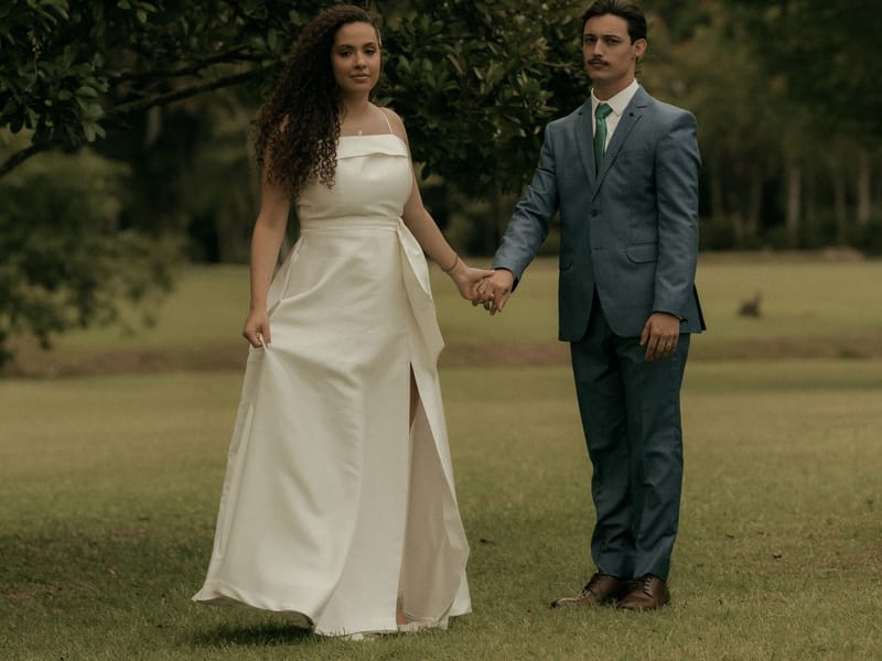 A bride and groom walking hand in hand across a lawn under a soft, overcast sky