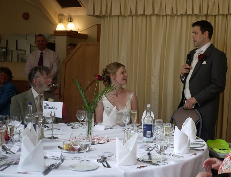 A groom giving his speech at the top table while the bride and her father listen